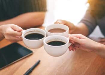 Top view image of three people clinking coffee cups on wooden table in cafe