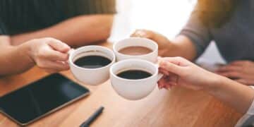 Top view image of three people clinking coffee cups on wooden table in cafe