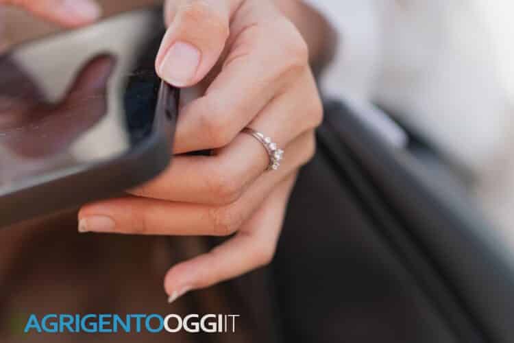 A close-up of a person's hand holding a smart phone, with a focus on the screen. The hand is adorned with a colorful bracelet, and the background is softly blurred, suggesting an outdoor setting.